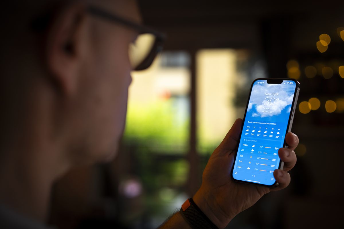 一名戴眼镜的男子看着手中的手机，屏幕上显示着苹果的天气应用程序。